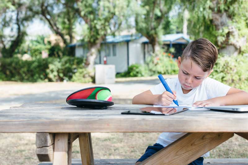 Un niño sentado en una mesa de picnic al aire libre, concentrándose en dibujar con marcadores, demostrando la importancia del estudio y el aprendizaje creativo. Sobre la mesa se coloca una bola de colores, con árboles y un edificio visibles al fondo.