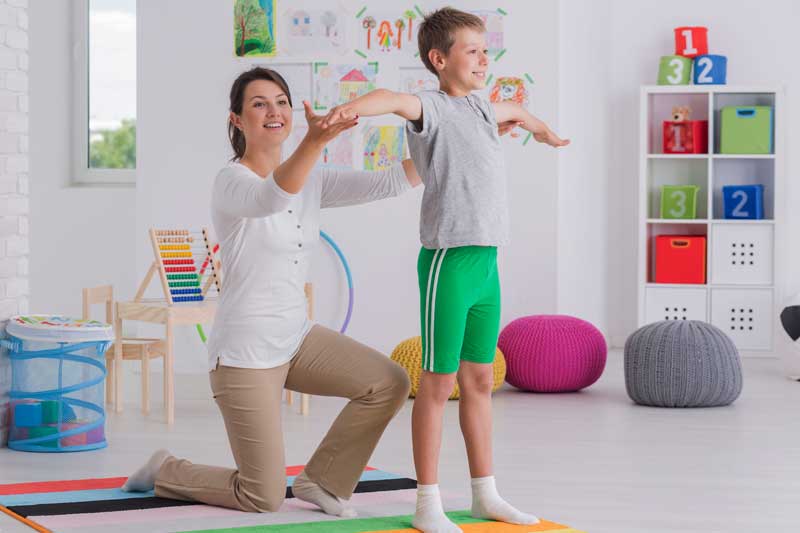 Una mujer se arrodilla detrás de un niño con los brazos extendidos en una sala de juegos interior llena de juguetes coloridos y arte en las paredes, creando un vibrante ámbito educativo.