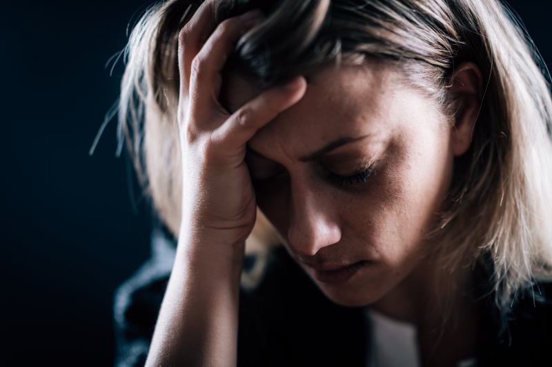 Una mujer de cabello rubio sostiene su cabeza con una mano, luciendo angustiada con los ojos cerrados, posiblemente abrumada por las emociones del confinamiento.