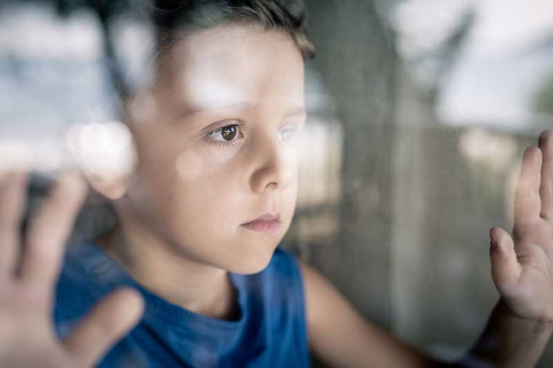 Un joven de pelo corto y camiseta azul sin mangas mira por una ventana con expresión pensativa, con las manos apoyadas en el cristal, mientras contempla la superación de su fracaso escolar.