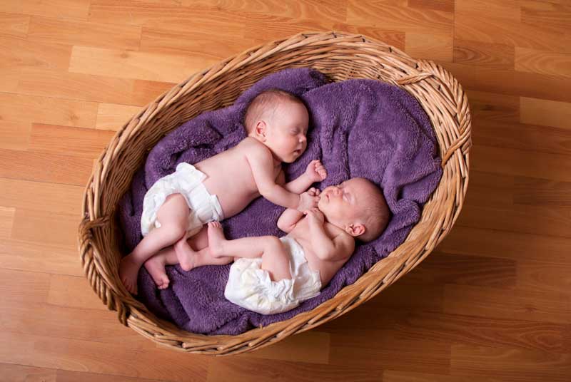 Dos bebés recién nacidos que usan pañales yacen dormidos en una canasta de mimbre forrada con una manta violeta, colocada sobre un piso de madera. La serena escena refleja las alegrías y los desafíos de los partos múltiples, recordando a los padres la importancia de la educación de hijos desde el principio.