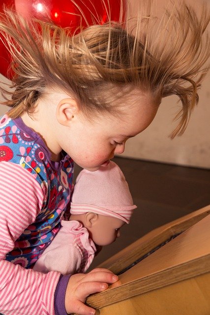 Un niño pequeño con el pelo alborotado mira fijamente un libro abierto mientras sostiene cerca una muñeca, capturando un momento de inocencia y concentración. Esta sentida escena se desarrolla en el Día Mundial del Síndrome de Down, celebrando la belleza de cada individuo.