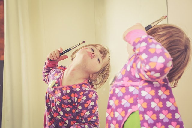 Un niño pequeño con Síndrome de Down con un pijama colorido con estampados de corazones sostiene una brocha de maquillaje e imita su reflejo en un espejo, celebrando su singularidad en el Día Mundial.