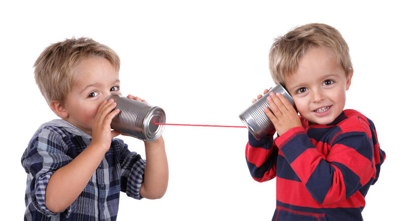 Dos niños pequeños juegan con un teléfono de lata, acercándose las latas a las orejas y sonriendo, conectados por un hilo rojo. Este sencillo juego no sólo les aportará alegría sino que también potenciará sus habilidades fonológicas.