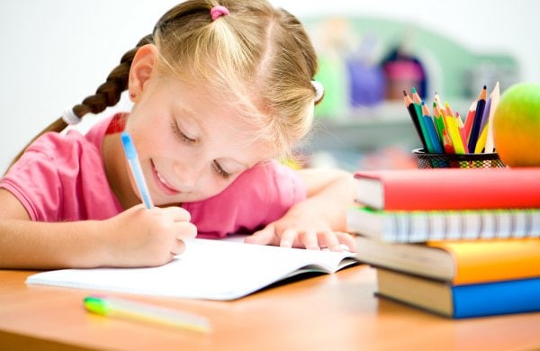 Una joven con cabello trenzado escribe en un cuaderno en un escritorio, mostrando su fuerte hábito de estudio. Hay libros coloridos y un contenedor de lápices de colores a su derecha.