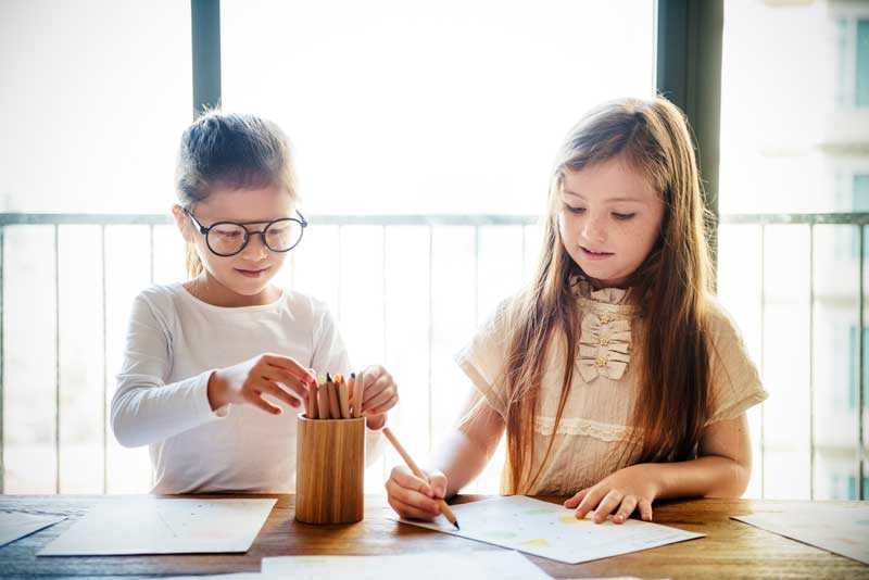 Dos niñas sentadas en una mesa dibujando con lápices de colores. Una niña usa gafas y selecciona un lápiz de un recipiente mientras la otra se concentra en su dibujo, distrayéndose momentáneamente de sus deberes.