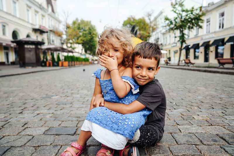 Dos niños sentados en una calle adoquinada; el niño mayor abraza a la niña más joven que se tapa la boca con una mano. Están rodeados de edificios y árboles, tal vez comunicándose en un lenguaje silencioso que sólo los hermanos entienden.