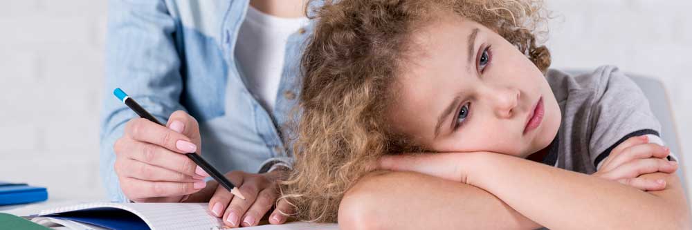 Un niño con cabello rizado apoya la cabeza sobre los brazos cruzados mientras está sentado en una mesa, luciendo cansado. Un adulto a su lado señala un cuaderno con un lápiz y ofrece orientación para ayudar al niño a superar los desafíos de Trastorno Pragmático.