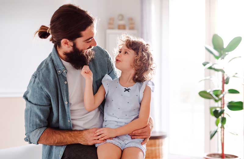 Un hombre con barba y un niño pequeño están sentados juntos en el interior, ambos sonriendo y mirándose. El fondo incluye una planta y una suave luz natural que entra por las ventanas. Parecen estar disfrutando de un momento de conexión, tal vez practicando técnicas de hablar para mejorar la comunicación entre ellos.