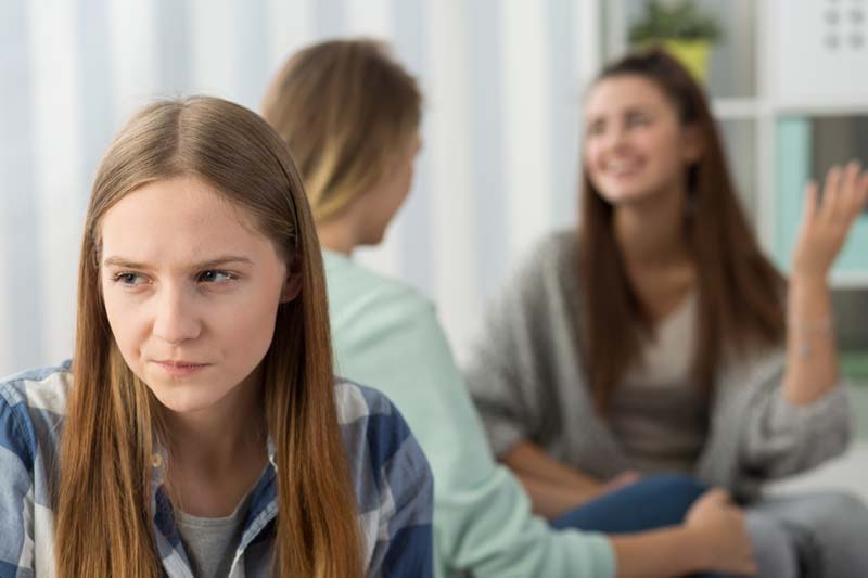 Tres mujeres jóvenes están en una habitación. Uno en primer plano parece molesto mientras los dos en el fondo están conversando, tal vez les vendría bien si pudieran comunicarse más.