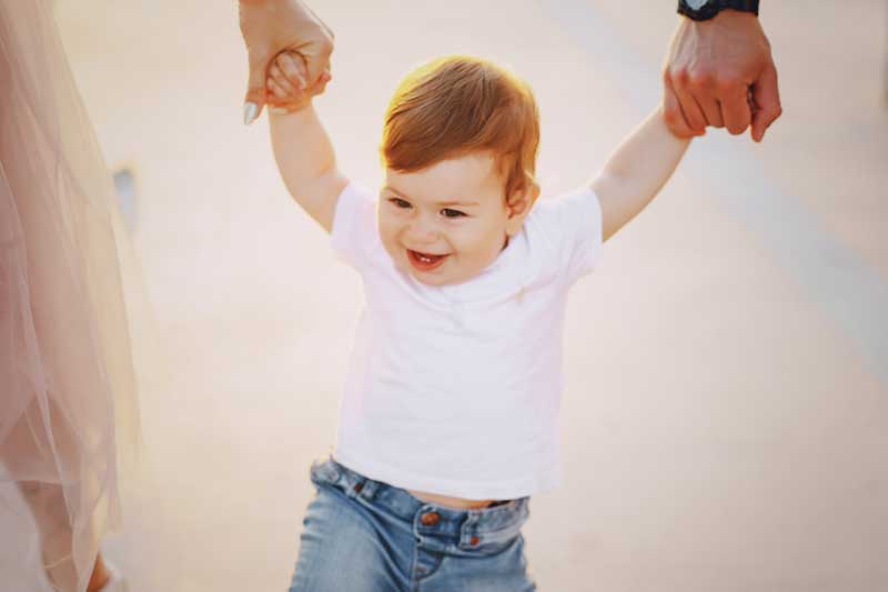 Un niño pelirrojo con camisa blanca y jeans es sostenido de las manos por dos adultos mientras camina, perfecto para hablar con nuestros hijos durante estos preciosos 12 a 18 meses.