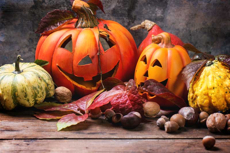 Dos calabazas talladas con caras felices, un toque de alegría de Halloween, rodeadas de calabazas variadas, coloridas hojas de otoño y nueces sobre una superficie de madera.