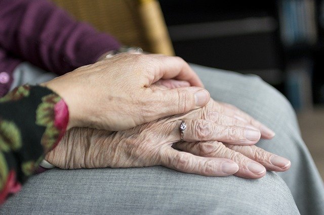 Se muestran dos manos apoyadas en la mano de otra persona. Una mano parece mayor con un anillo y la otra, posiblemente afectada por la enfermedad de Parkinson, está colocada en la parte superior. El fondo está borroso.