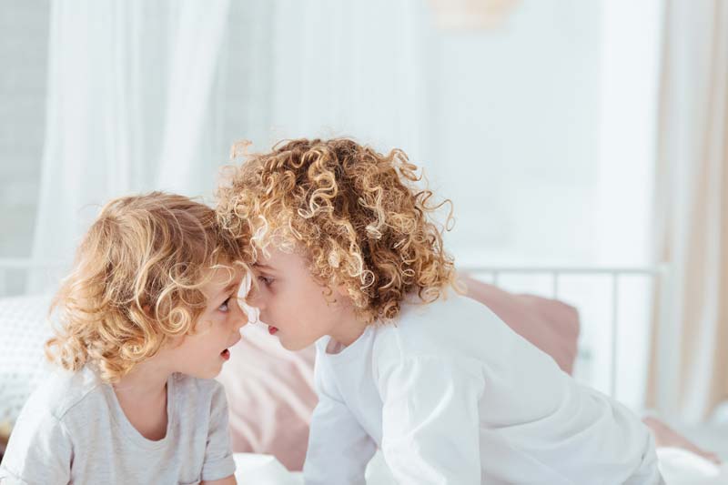 Dos niños pequeños de pelo rizado, tal vez después de una de sus habituales peleas entre hermanos, se tocan la frente y se miran con ternura, sentados en una cama en una habitación suavemente iluminada.