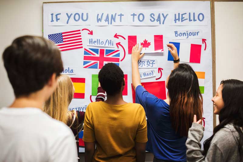 Un grupo de cinco personas se para frente a un mural que muestra cómo decir "Hola" en varios idiomas, con las banderas de los países correspondientes, promoviendo el aprendizaje de idiomas para sus hijos.