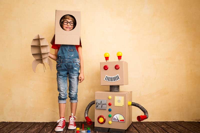 Un niño, lleno de ilusión, lleva un casco de cartón y sostiene un cohete de cartón, de pie junto a un robot de cartón casero con varios controles dibujados. Esta escena captura las ganas de hacer cosas que realmente despiertan creatividad y alegría en las mentes jóvenes.