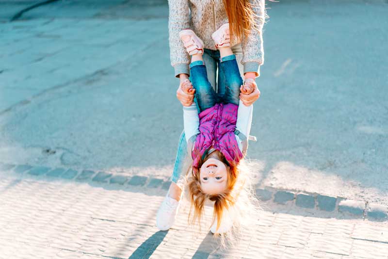 Un adulto sostiene con las manos a un niño sonriente boca abajo en un día soleado en una obra de teatro inversa. El niño lleva un chaleco rosa y jeans azules mientras mira juguetonamente a la cámara.