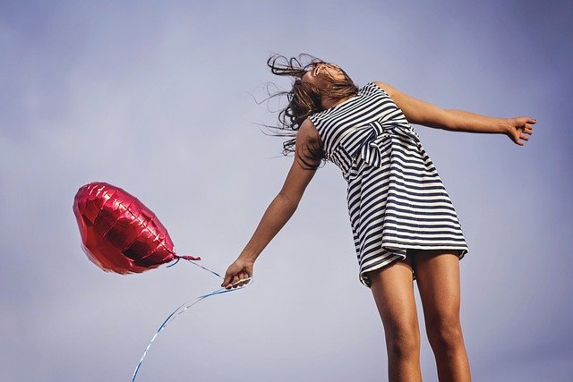 Una niña con un vestido a rayas sostiene un globo rojo en forma de corazón y parece estar saltando contra un fondo de cielo azul, capturando el espíritu de FELICIDAD. Este momento de alegría es perfecto para celebrar el DÍA INTERNACIONAL DE LA FELICIDAD el 20 DE MARZO.