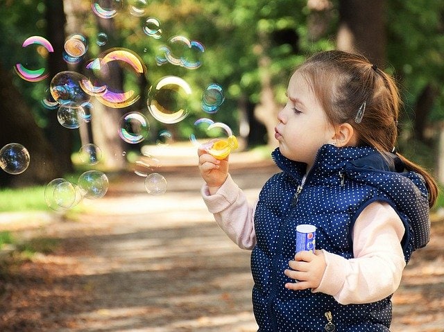 Un niño pequeño hace pompas de jabón al aire libre en un camino rodeado de árboles, disfrutando de los beneficios simples del juego mientras sostiene una varita y un recipiente para hacer burbujas, lo que recuerda a la terapia orofacial en su alegría y valor de desarrollo.