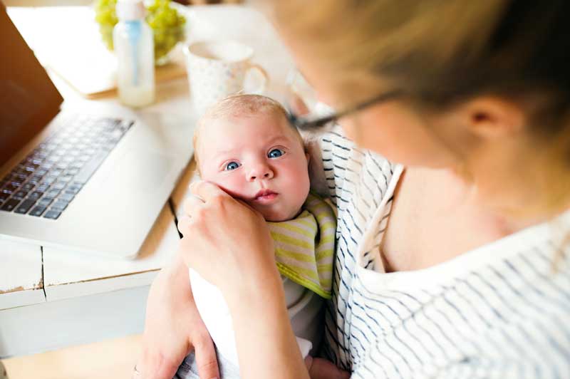 Una persona que sostiene a un bebé mientras está sentada en un escritorio con una computadora portátil y un biberón cerca, ofreciendo apoyo esencial en lactancia materna para niños prematuros.