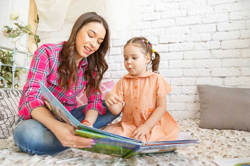 Una mujer y una niña sentadas juntas en una cama, profundamente ocupadas en lectura y escritura. La mujer, vestida con una camisa a cuadros, y la niña con un vestido naranja están absortas en el libro, compartiendo su amor por la lectura.