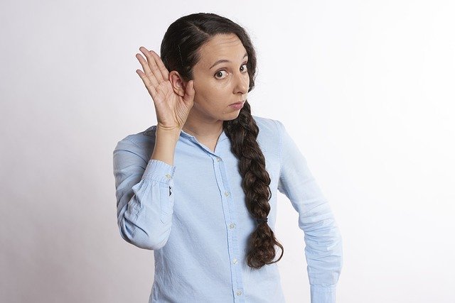 Una persona con el pelo largo trenzado y una camisa azul claro se lleva una mano a la oreja, tal vez experimentando cierta pérdida de audición mientras intenta escuchar con atención.