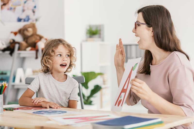 Un niño y un adulto se sientan en una mesa con materiales de arte y aprendizaje; el adulto sostiene un papel con una gran letra A roja mientras involucra al niño, utilizando herramientas logopedia para mejorar su sesión interactiva.