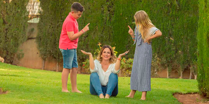 Una mujer sentada en el césped con los brazos en alto participa en un programa "ser una mejor madre", mientras un niño y una niña, ambos apuntándola con el dedo emocionados, se encuentran cerca en un jardín.