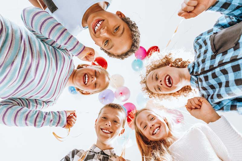 Un grupo de cinco niños sonriendo y mirando a la cámara con globos de colores al fondo. Parecen felices y están al aire libre en un día soleado, disfrutando de lo que parece una tarde alegre después de una divertida sesión de terapia en grupo.