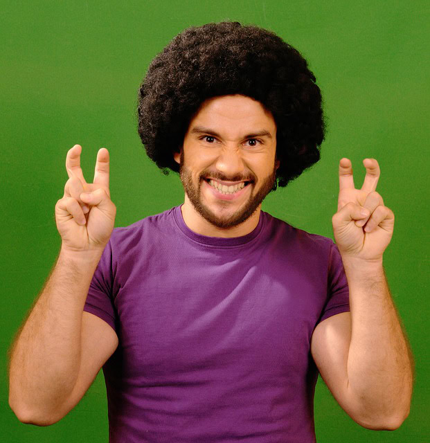 Un hombre sonriente con peinado afro y camiseta morada hace comillas en el aire con ambas manos sobre un fondo verde, destacando la importancia de la comunicación no verbal.