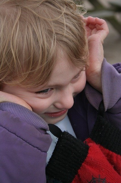 Un niño pequeño de cabello rubio que viste una chaqueta morada y se sujeta las orejas, sonríe y mira hacia abajo, mostrando resiliencia en su manejo emocional.