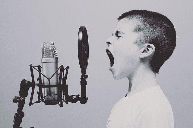 Un niño está de perfil, gritando o cantando apasionadamente frente a un micrófono con un filtro pop en un estudio, tal vez preparándose para las festividades de la "vuelta al cole".