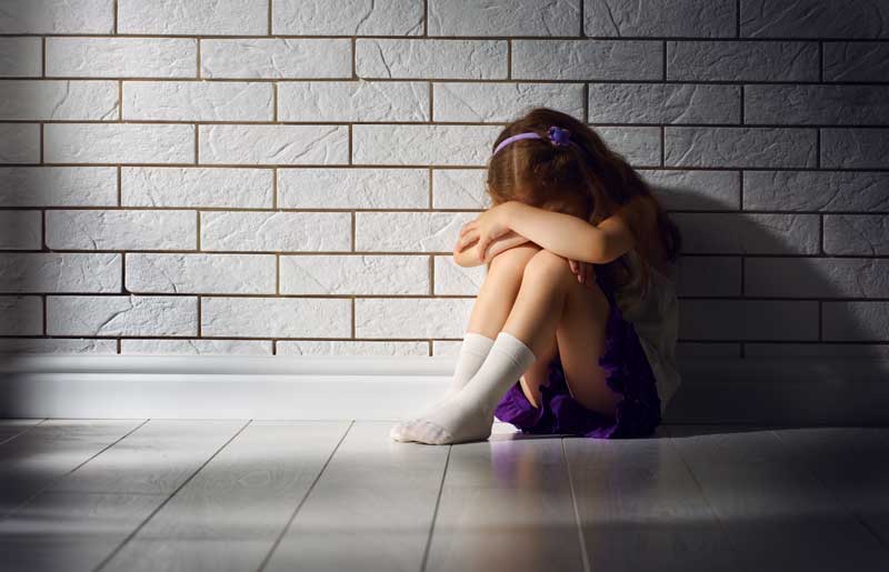 Una niña sentada en el suelo contra una pared de ladrillos blancos, abrazándose las rodillas y ocultando su rostro. Lleva una camisa blanca, falda morada, calcetines blancos y un accesorio para el cabello morado. Consumida por los miedos, tal vez necesite que alguien la ayude en este momento.