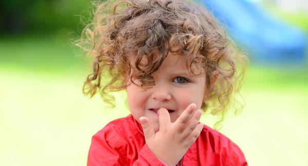 Un niño pequeño con cabello rizado, probablemente un *hijo*, vestido con una chaqueta roja, se toca la boca con una mano mientras está parado al aire libre.