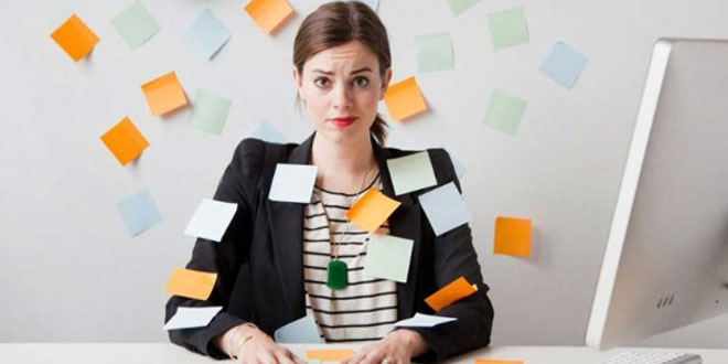 Una mujer sentada ante un escritorio cubierta de notas adhesivas, parece abrumada. Está frente a una computadora, con más notas adhesivas en la pared detrás de ella, profundamente inmersa en su cultura de ocupación diligente.