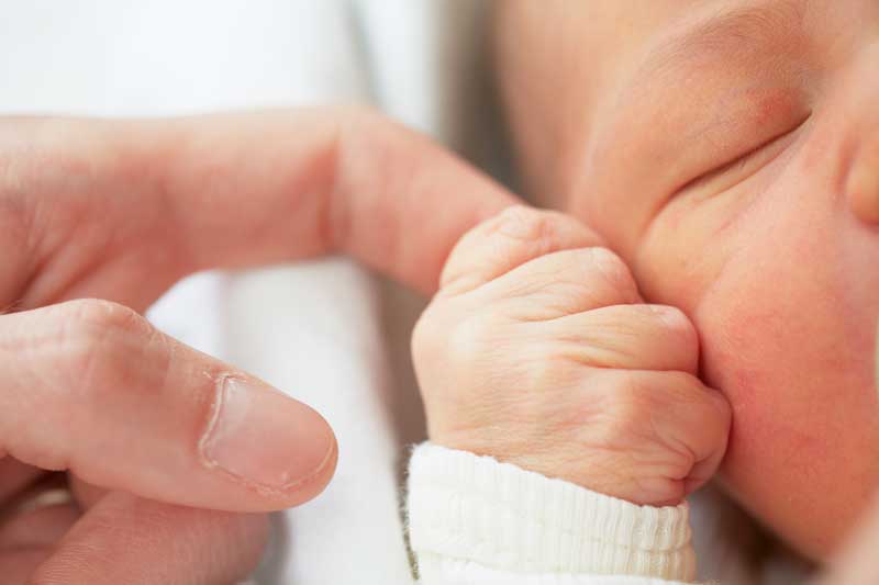 Primer plano de un recién nacido, nacido en diciembre, sosteniendo el dedo de un adulto mientras duerme. Sólo se ve una parte del rostro del recién nacido. La mano del adulto toca suavemente la mano del recién nacido. Ambos parecen estar descansando.