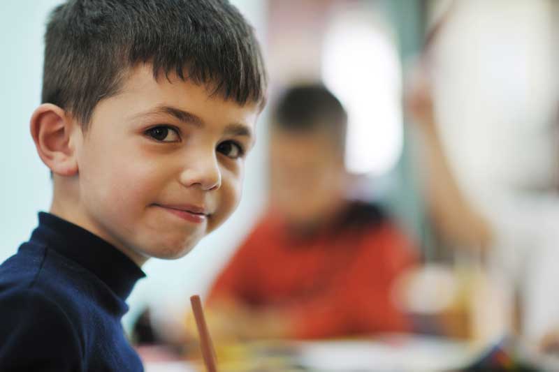 Un joven de cabello oscuro, vestido con una camisa oscura, mira a la cámara mientras está sentado en un salón de clases con otros niños en el fondo borroso. Parece pensativo, tal vez reflexionando sobre cómo superar su disfemia y hablar con confianza durante la clase.