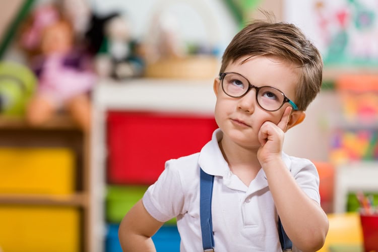 Un niño pequeño con cabello castaño corto y gafas, vestido con una camisa blanca y tirantes azules, permanece en el interior durante el encierro con una expresión pensativa, tocándose la mejilla con un dedo.