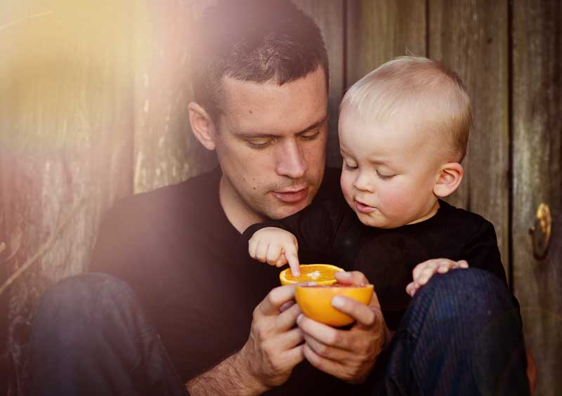 Un hombre y un niño pequeño están sentados juntos sobre un fondo de madera, ambos con camisas negras. El niño señala un cuenco naranja que sostiene el hombre, generando un momento de unión y potencialmente ayudando en el desarrollo del habla.