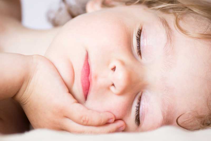 Un primer plano de un niño dormido apoyando la cabeza en la mano. Los ojos del niño están cerrados y los labios ligeramente entreabiertos, captando la tranquilidad que se obtiene al fomentar hábitos saludables en nuestros hijos.