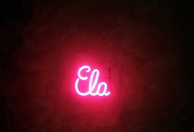 Un letrero de neón con la palabra "ELA" en cursiva, brillando en rosa sobre un fondo oscuro.