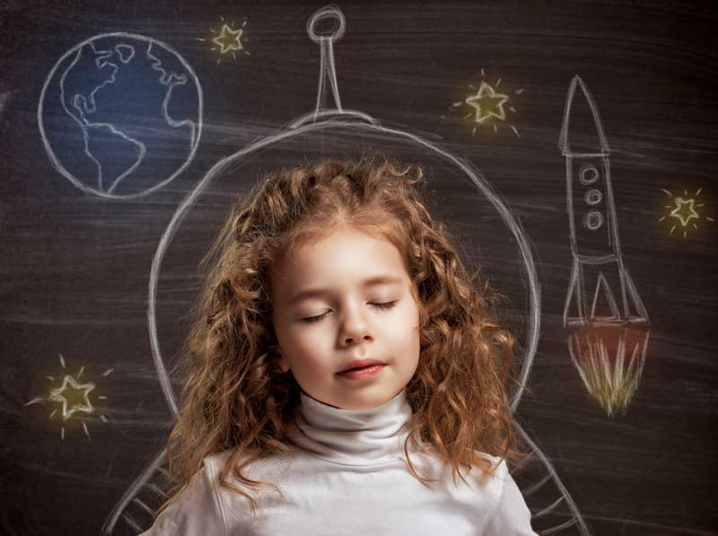 Un niño con cabello rizado y ojos cerrados se para frente a un dibujo en pizarra de una nave espacial, las estrellas y la Tierra, encarnando los sueños que tenemos para nuestros hijos mientras aprenden a respetar las normas mientras exploran su imaginación.