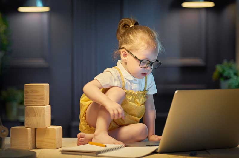 Un niño pequeño con gafas se sienta en una mesa, explorando nuevas tecnologías en una computadora portátil. Sobre la mesa también hay bloques de madera y un cuaderno con un lápiz, como si estuvieran listos para capturar la magia del aprendizaje de idiomas.