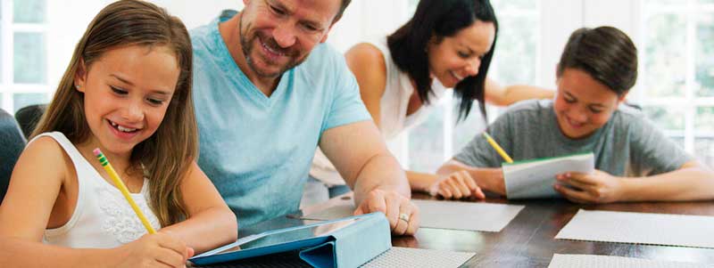 Una familia sentada junta en una mesa, con el padre y la hija trabajando en una tableta, mientras la madre y el hijo escriben en cuadernos, resaltando la importancia de la educación de los hijos y la unión de los padres.