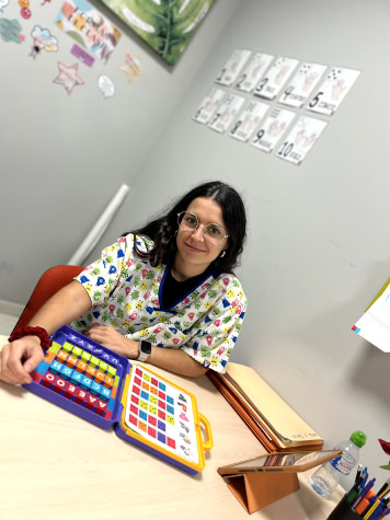 Una mujer con gafas se sienta en una mesa con un colorido juego de mesa frente a ella. Las paredes circundantes tienen carteles educativos y artículos de Nosotras.