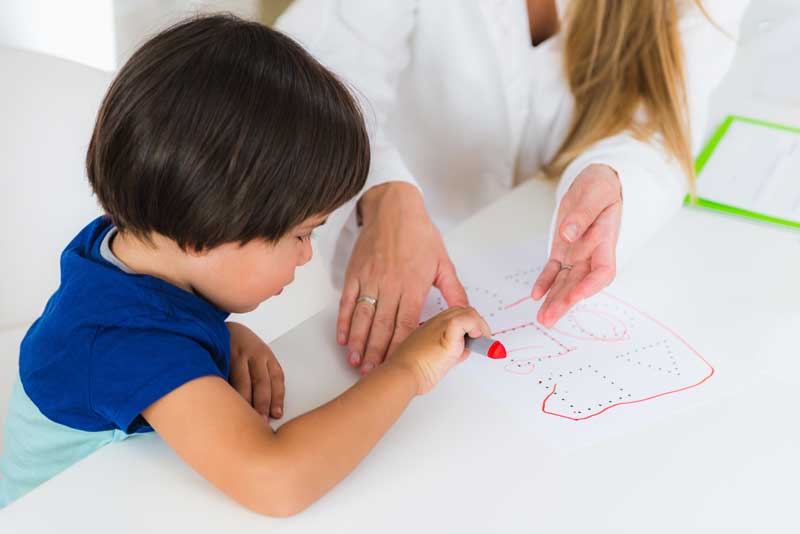 Un niño, ayudado por un adulto, utiliza un crayón rojo para conectar puntos en una hoja de papel que forma un dibujo, una actividad que incorpora sutilmente principios de la psicología para mejorar sus habilidades de desarrollo.