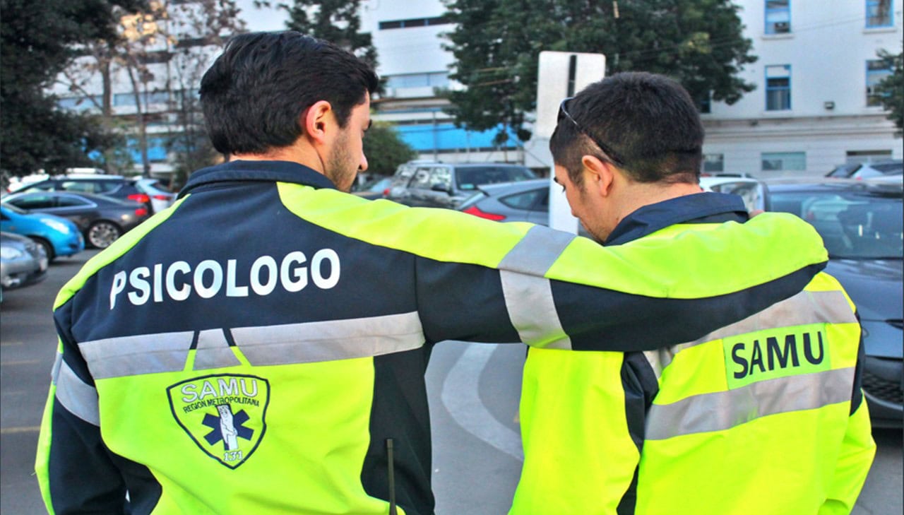 Dos hombres con chaquetas de alta visibilidad con las palabras "PSICOLOGO" y "SAMU" están de pie con el brazo de un hombre alrededor del otro, posiblemente brindando apoyo para un traumatismo indirecto. Parecen estar en una zona de aparcamiento al aire libre.