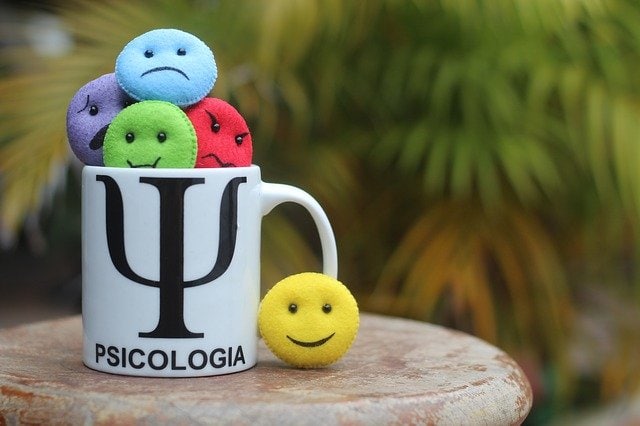 Una taza con la etiqueta "PSICOLOGIA" con un símbolo de psicología contiene emoticonos que expresan diferentes emociones: cara triste azul, cara preocupada morada, cara enojada roja, cara feliz verde y cara sonriente amarilla. Perfecto para recordarnos los diversos sentimientos que encontramos a diario.