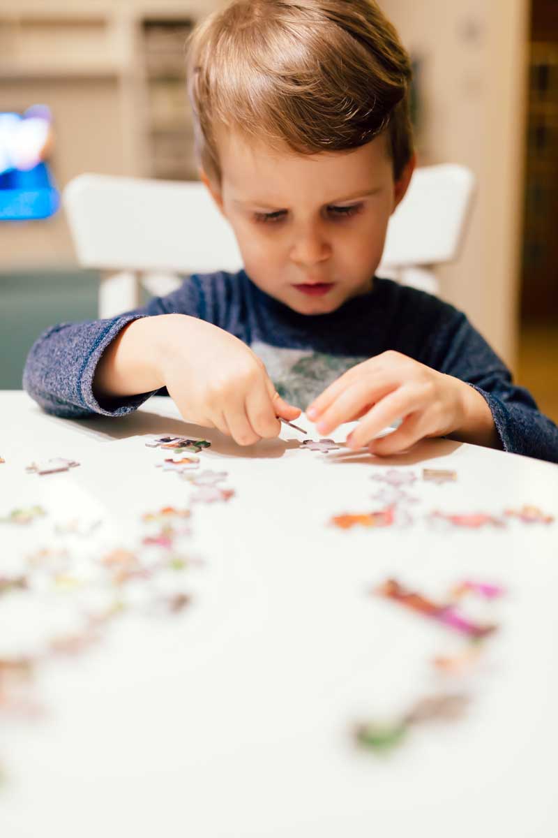 Un niño pequeño con un suéter azul se concentra en ensamblar las piezas de un rompecabezas en una mesa blanca en el interior, lo que ejemplifica la concentración silenciosa incluso mientras maneja los desafíos del trastorno del aprendizaje no verbal (TANV).