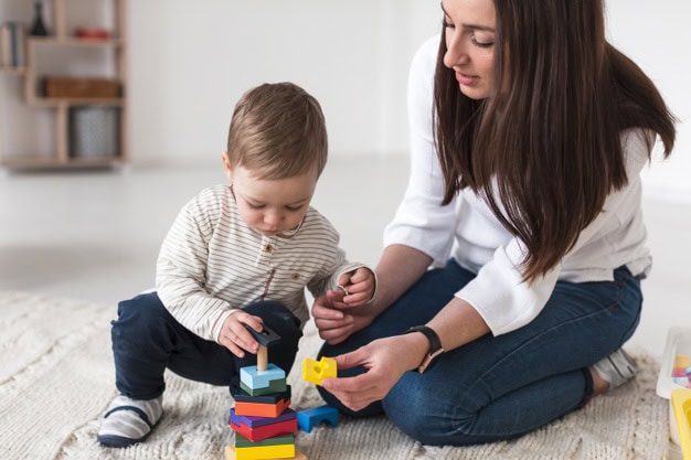 Un adulto y un niño están sentados en el suelo, en casa. El niño apila bloques de colores mientras el adulto sostiene un bloque y observa, un juego atractivo que puede mejorar el lenguaje de los hijos.